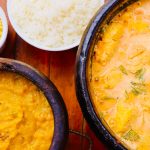 Moqueca brasileira colorida servida em uma panela, acompanhada de outros pratos sobre uma mesa de madeira.