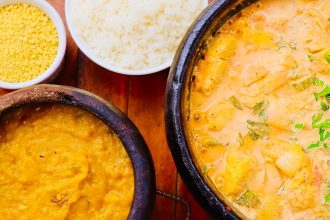 Moqueca brasileira colorida servida em uma panela, acompanhada de outros pratos sobre uma mesa de madeira.