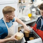 Dois funcionários preparam pedidos para entrega em uma pequena cozinha comercial.