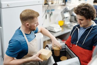 Dois funcionários preparam pedidos para entrega em uma pequena cozinha comercial.