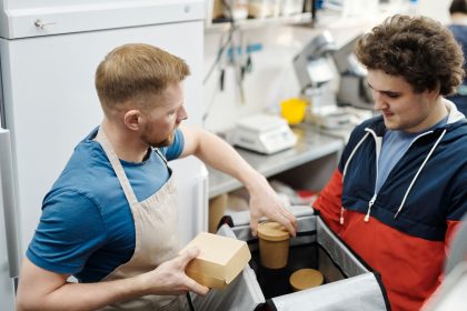 Dois funcionários preparam pedidos para entrega em uma pequena cozinha comercial.